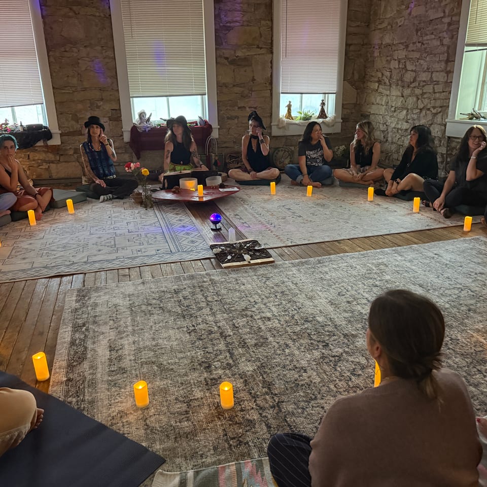 Group of people sit in a circle amid rugs and candles inside a warmly lit stone-walled room.