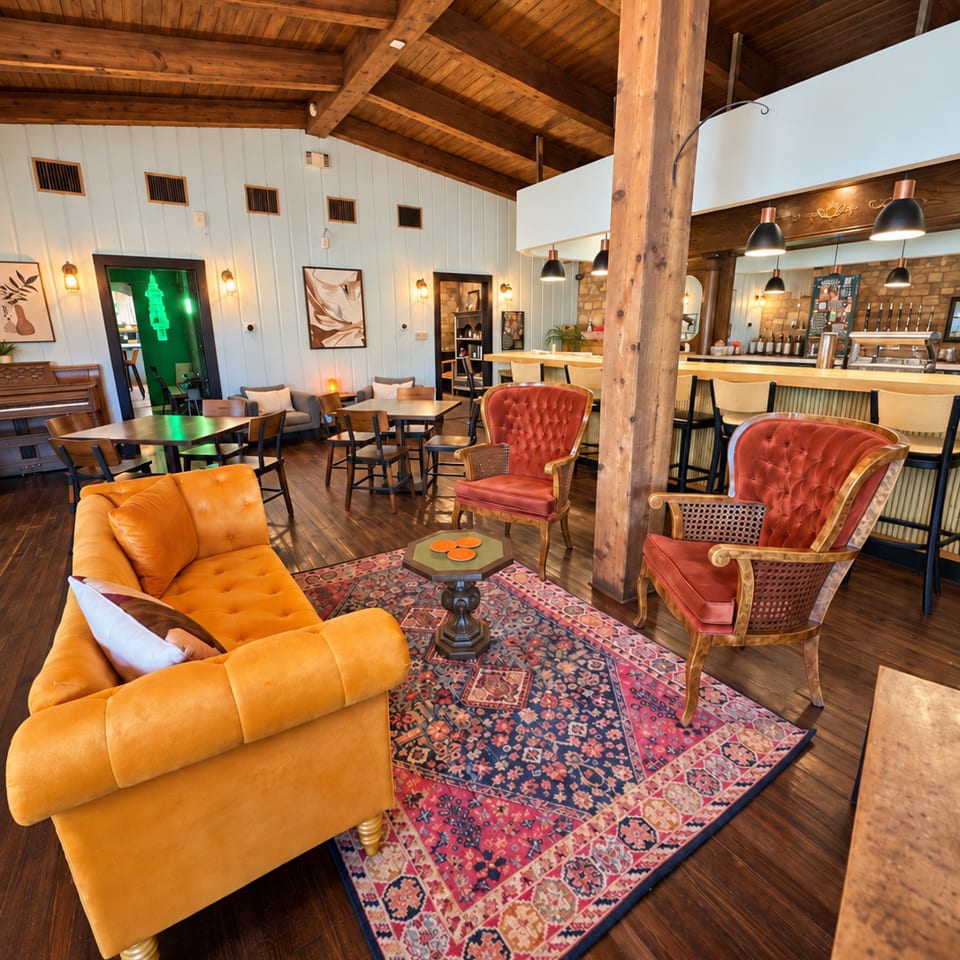 Rustic restaurant lounge with exposed wood beams, colorful vintage seating, patterned rug, bar, and warmly lit dining area.
