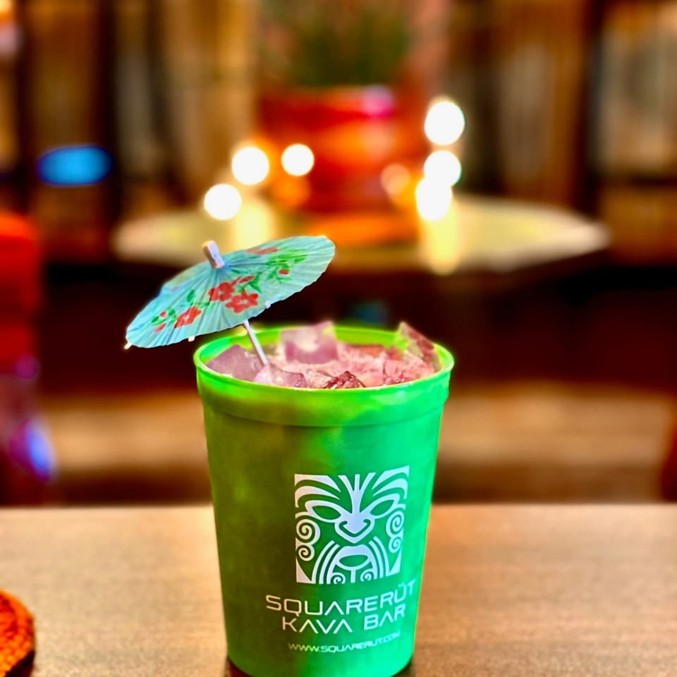 Bright green Square Root Kava Bar drink topped with ice and a floral paper parasol on a warmly lit bar counter.