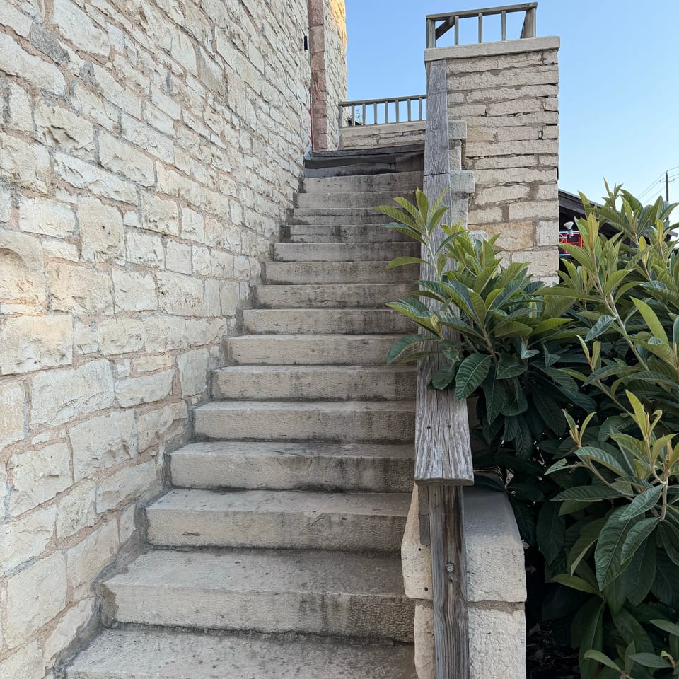 Stone staircase with wooden railings beside a rustic stone building, surrounded by leafy plants under a clear blue sky.