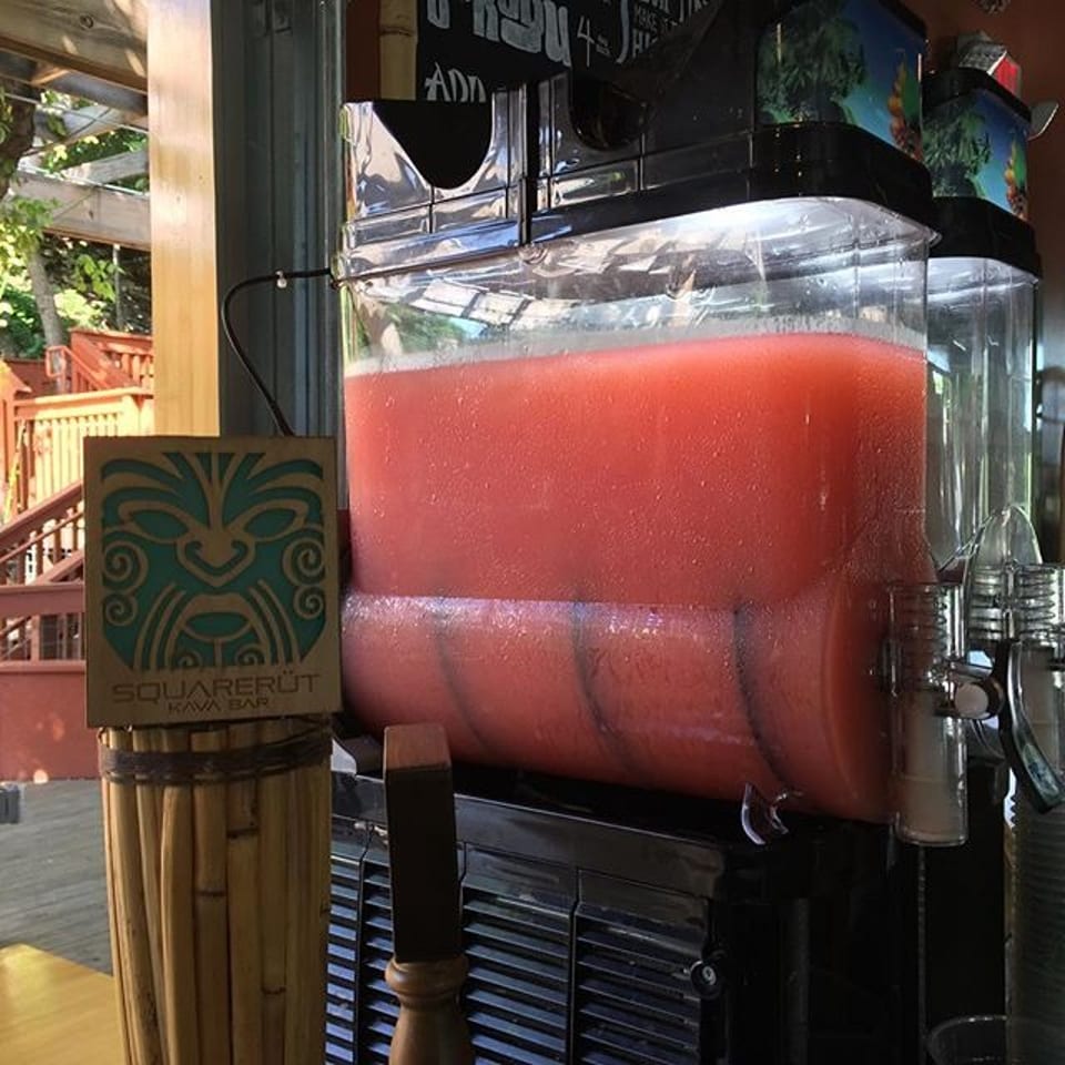 Large beverage dispenser filled with red frozen drink beside a Squareroot Kava Bar sign on a tropical outdoor patio.