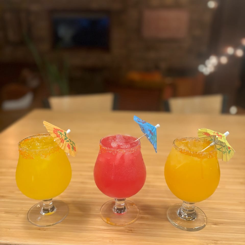 Three colorful cocktails with salted rims, ice, straws, and paper umbrellas sit on a wooden restaurant table.