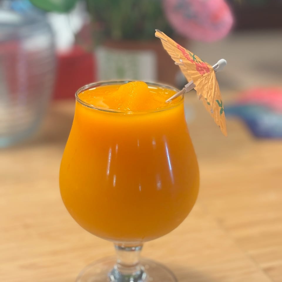 Bright orange cocktail in a stemmed glass, garnished with an orange slice and decorative paper umbrella on a restaurant table.