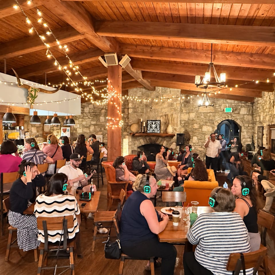 Busy rustic restaurant dining room with stone walls, wooden beams, string lights, fireplace, bar, and groups of patrons.