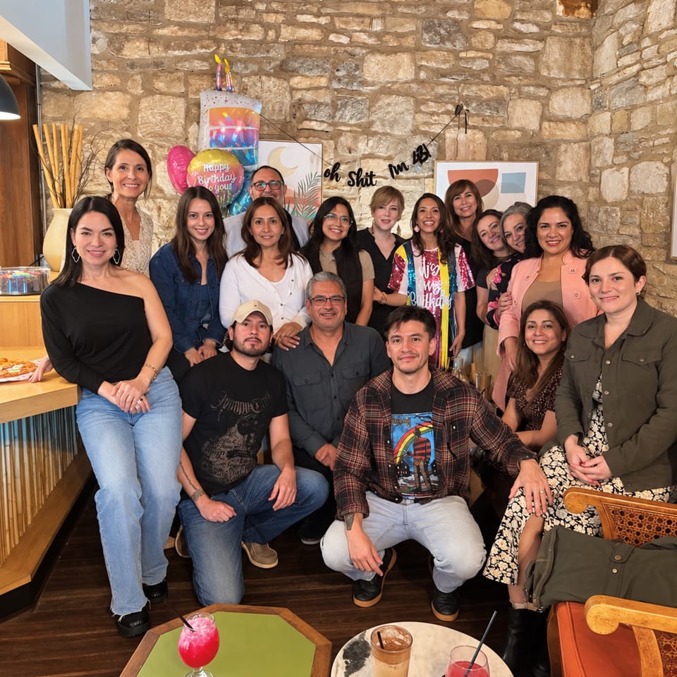 Group celebrating a birthday inside a rustic restaurant, gathered beneath balloons and a birthday banner around tables with colorful drinks.