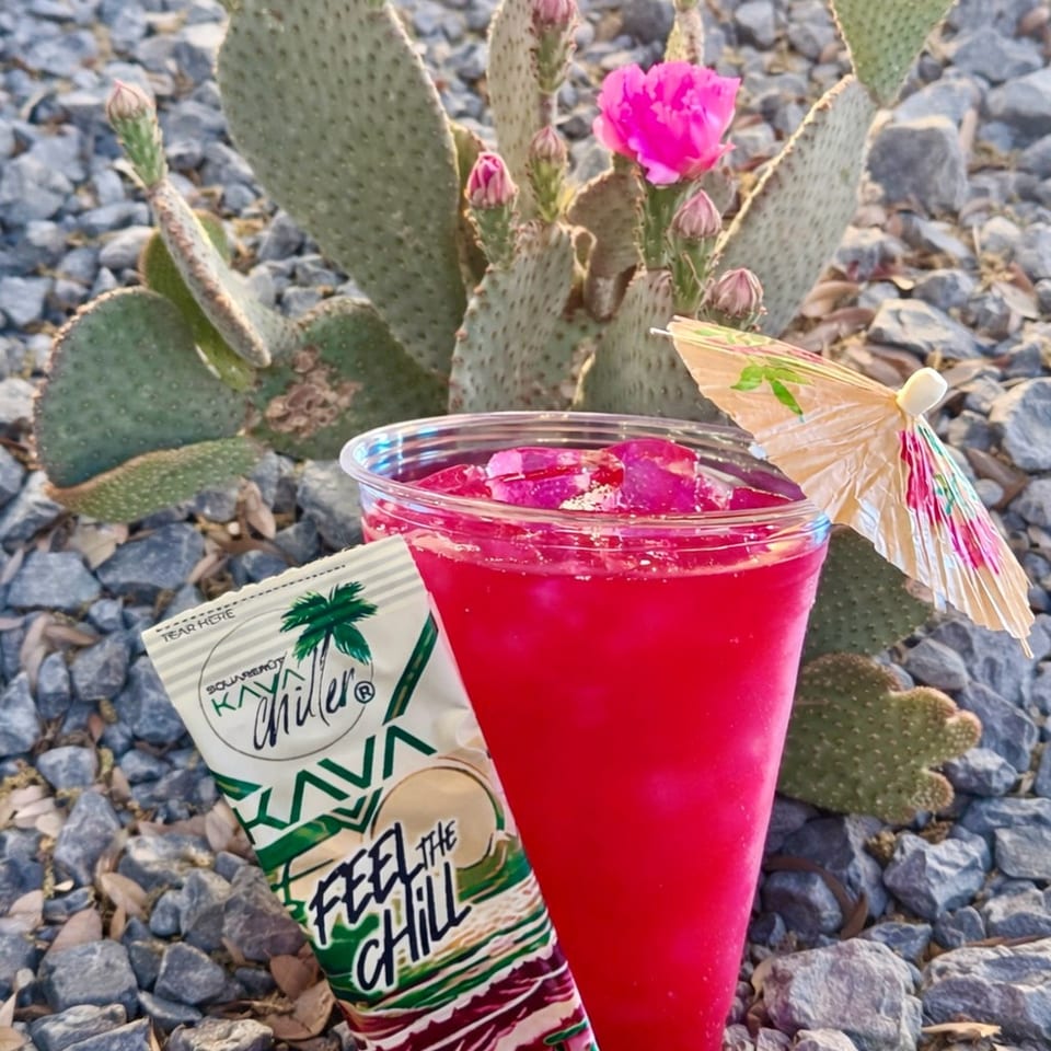 Bright red frozen drink with a cocktail umbrella beside a colorful chili-themed snack package, photographed in front of flowering prickly pear cactus.