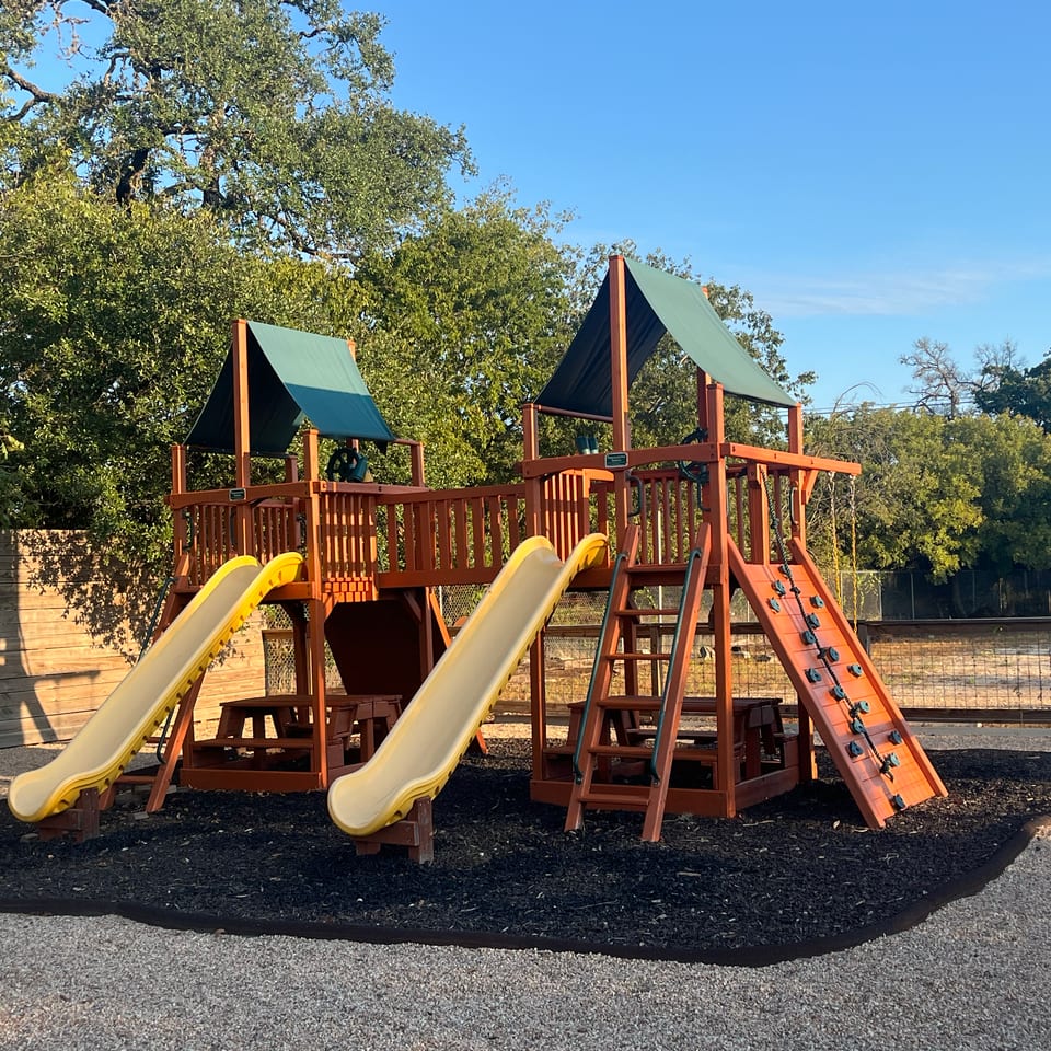 Outdoor wooden playground with yellow slides, green shade canopies, climbing wall, and surrounding trees.