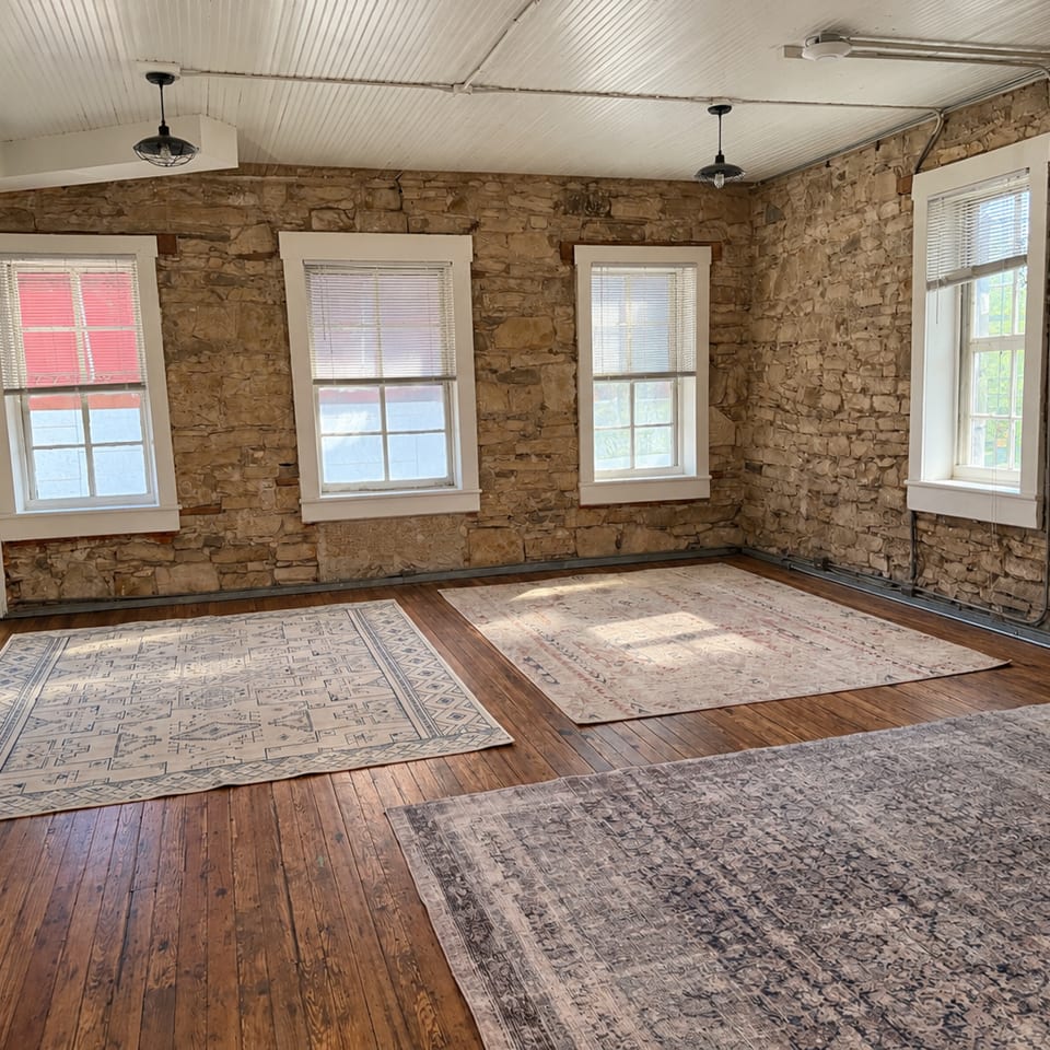 Rustic restaurant room with exposed stone walls, hardwood floors, four windows, pendant lights, and three patterned area rugs.
