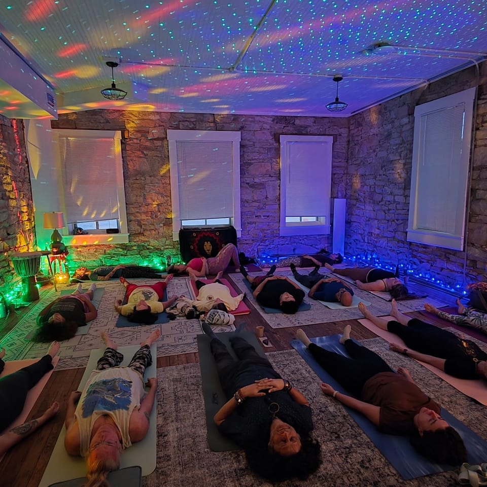 People lie on yoga mats in a rustic stone room illuminated by colorful disco lights.