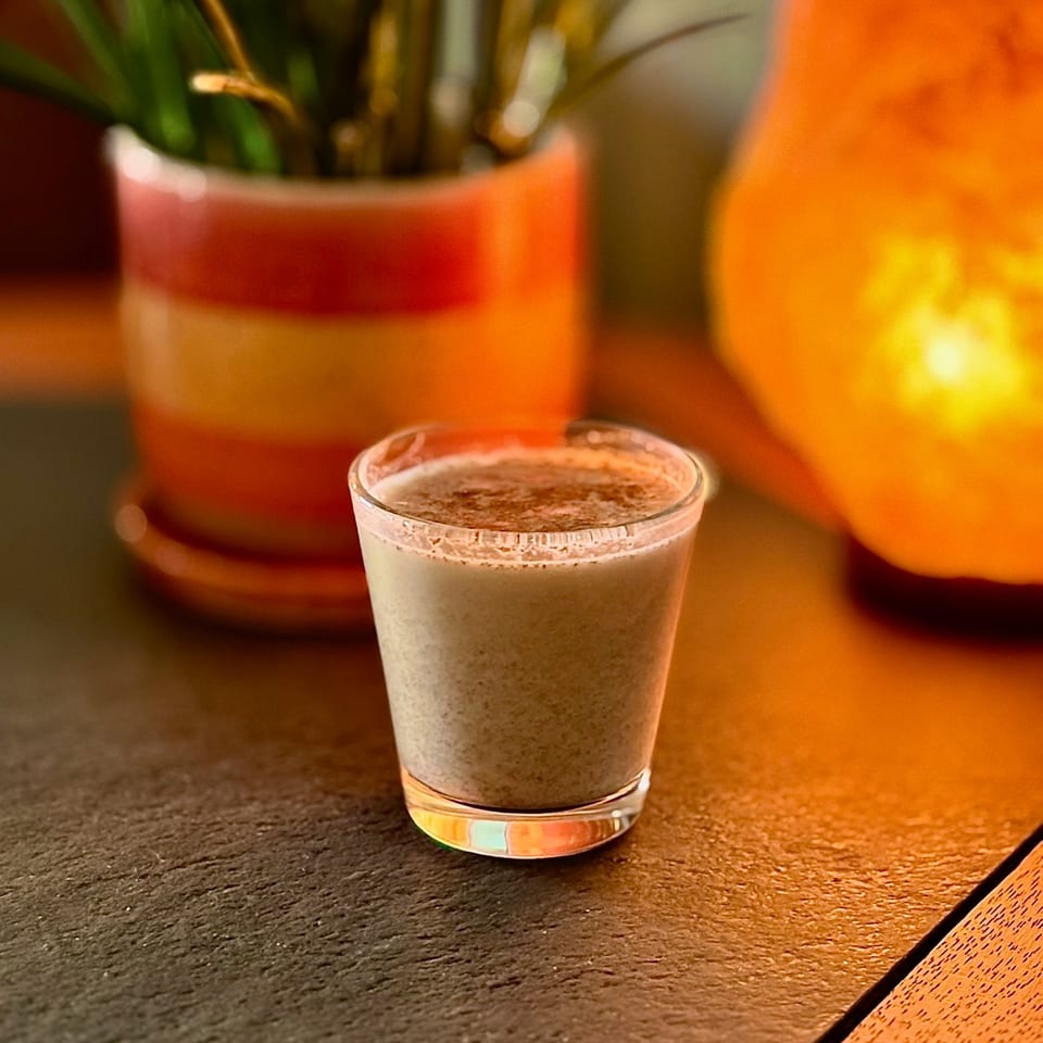 A creamy cocktail in a short glass sits on a dark table, warmly lit beside a potted plant and glowing orange lamp.