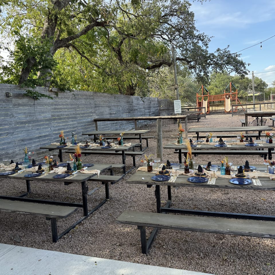 Outdoor restaurant patio with decorated picnic tables, string lights, mature trees, and a children’s playground.