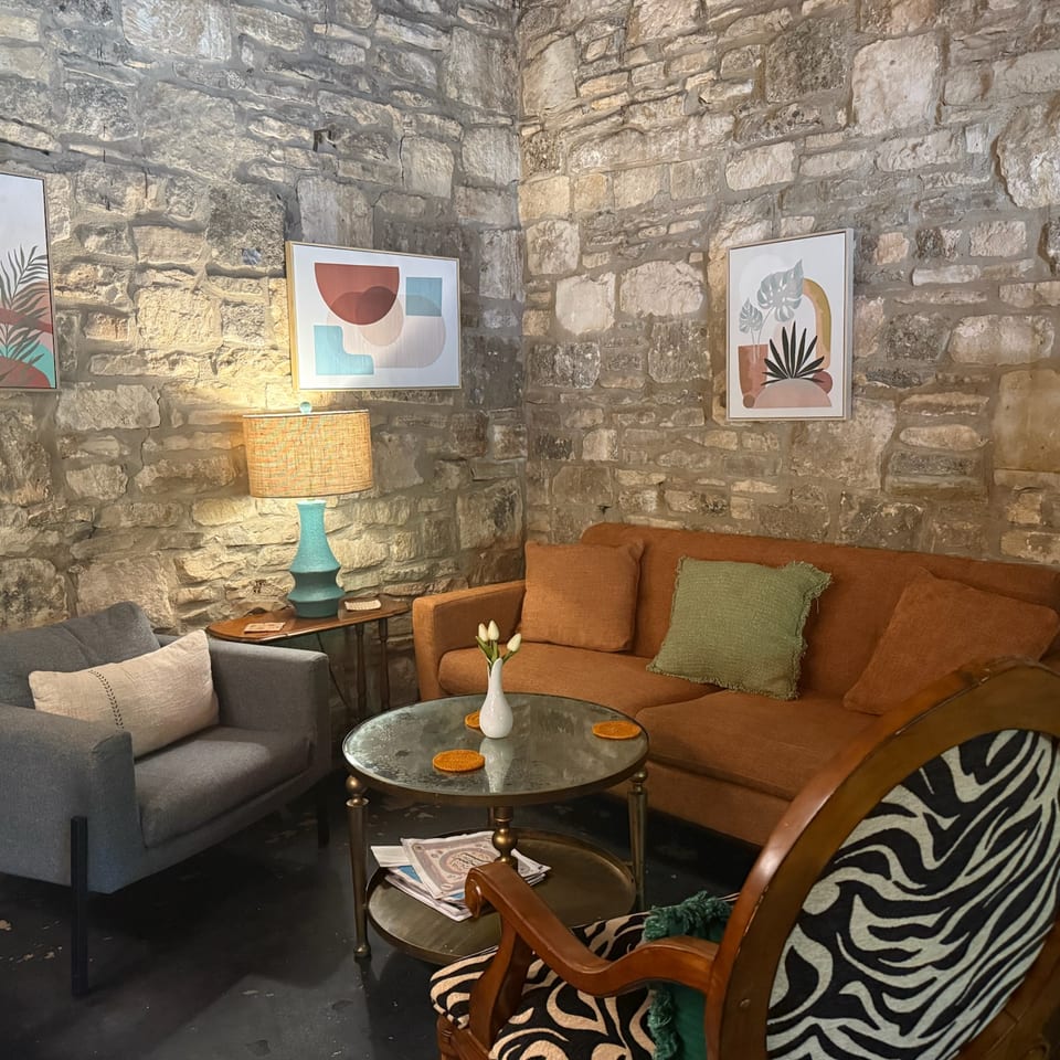Cozy restaurant lounge with exposed stone walls, colorful artwork, armchairs, rust-colored sofa, coffee table, and warm lamp lighting.