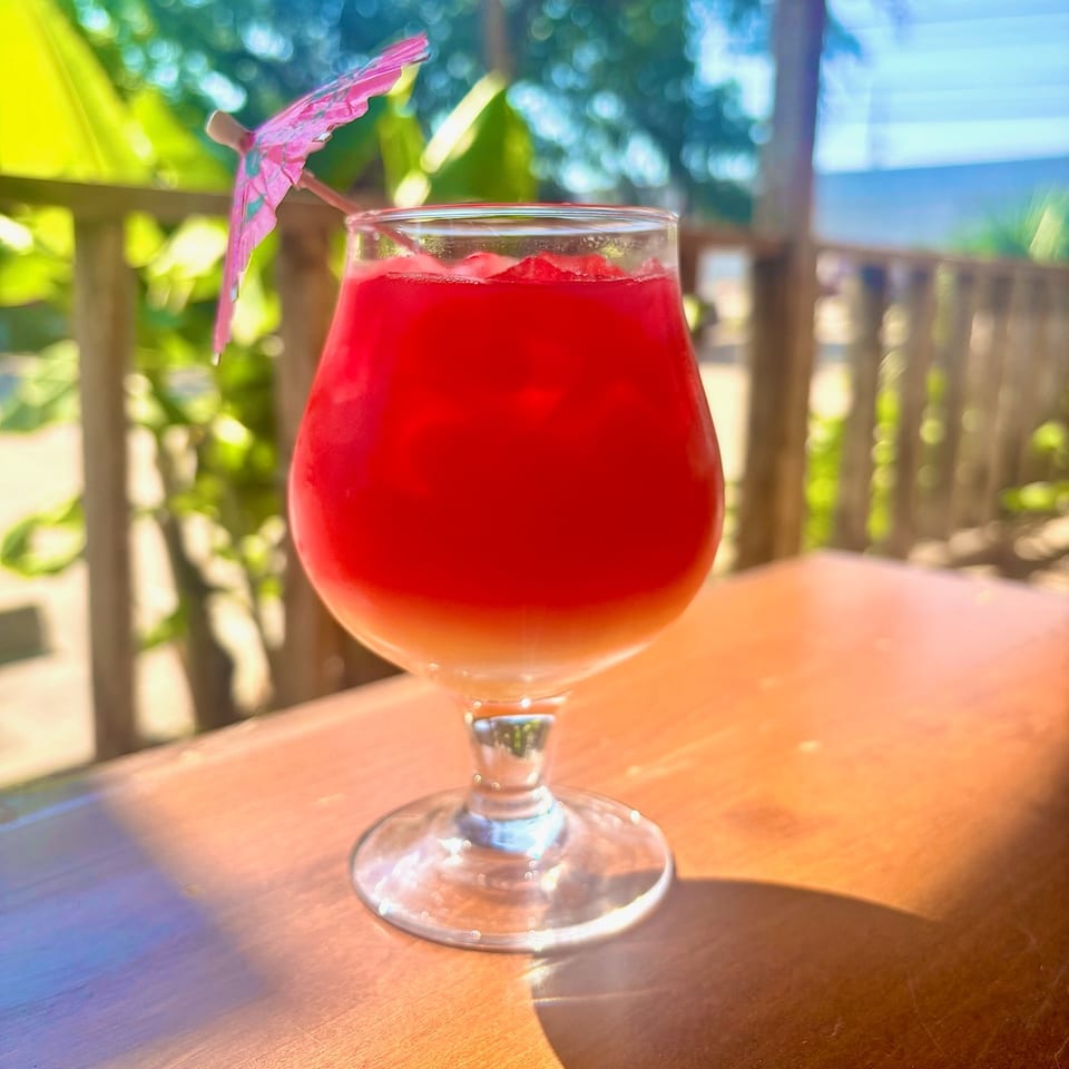 Bright red iced cocktail in a stemmed glass garnished with a pink paper umbrella on a sunlit outdoor table.