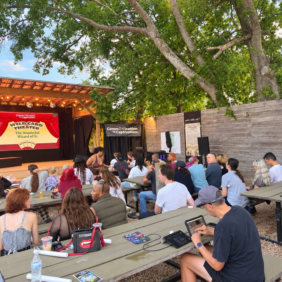 Outdoor restaurant theater with picnic tables, a Wizard of Oz stage, string lights, and a seated audience under leafy trees.