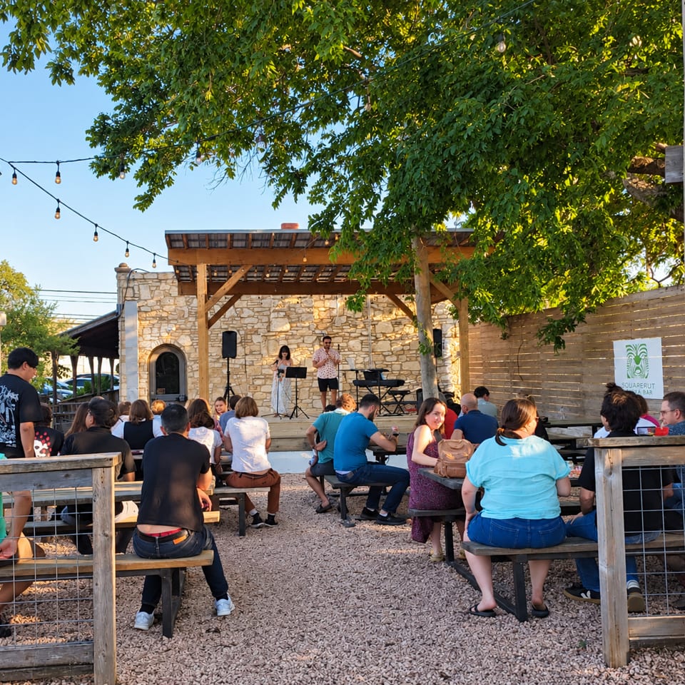 Outdoor restaurant patio with string lights, stone wall, live musicians performing, and guests seated at picnic tables.