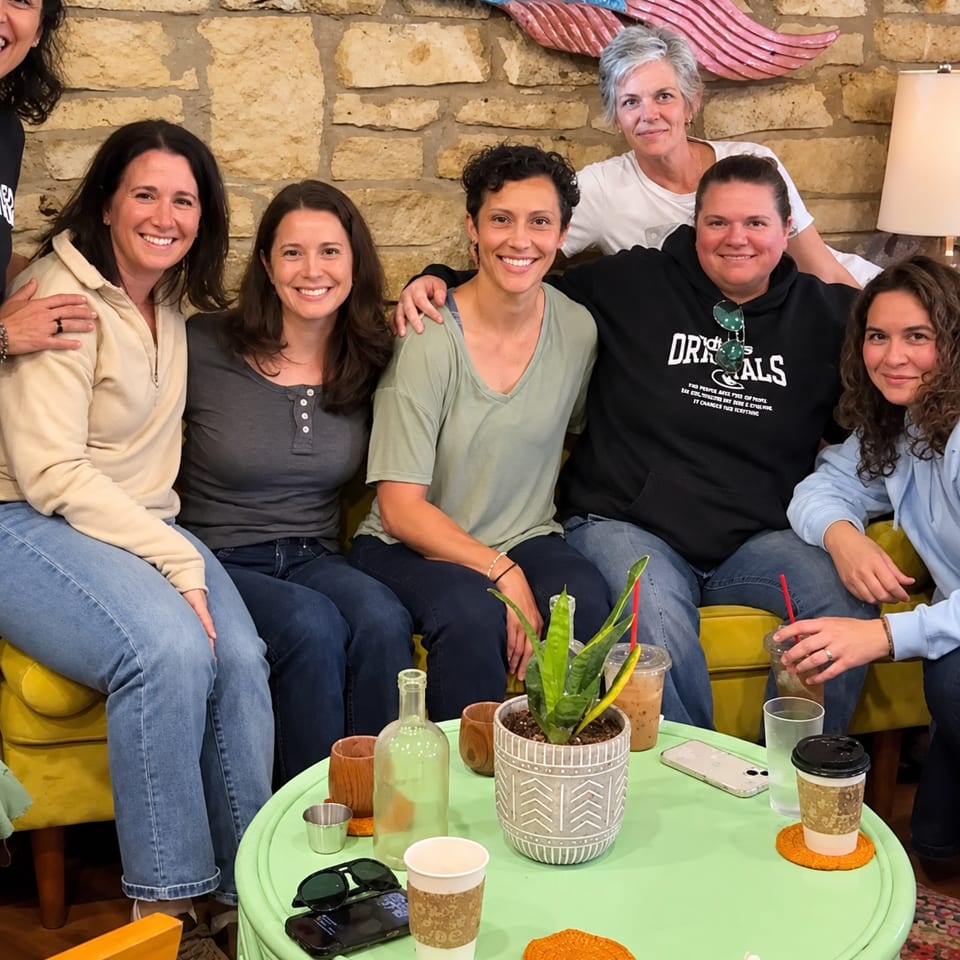 Smiling group gathered around a green café table with drinks and a potted plant against a stone wall.