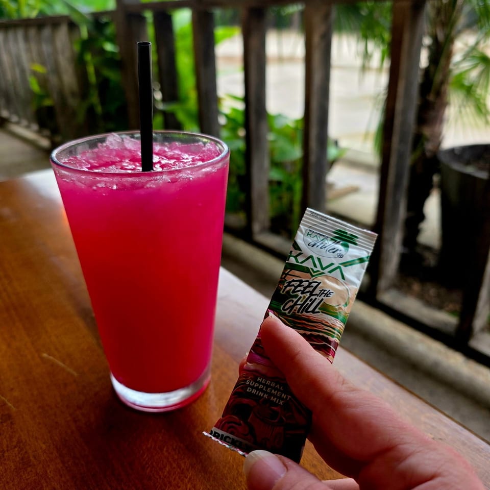 Bright pink iced drink with a black straw beside a hand holding a herbal supplement packet on a wooden table overlooking tropical greenery.